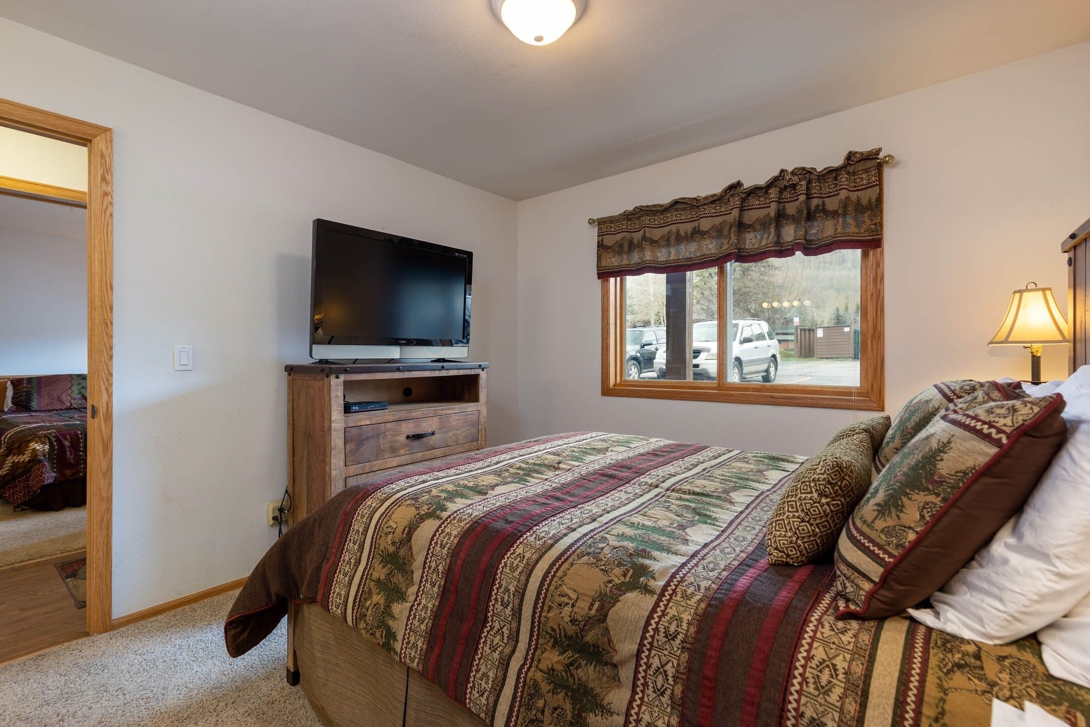 A bedroom with a bed, a television on a wooden stand, a window with a decorative valance, and a lamp on a bedside table.