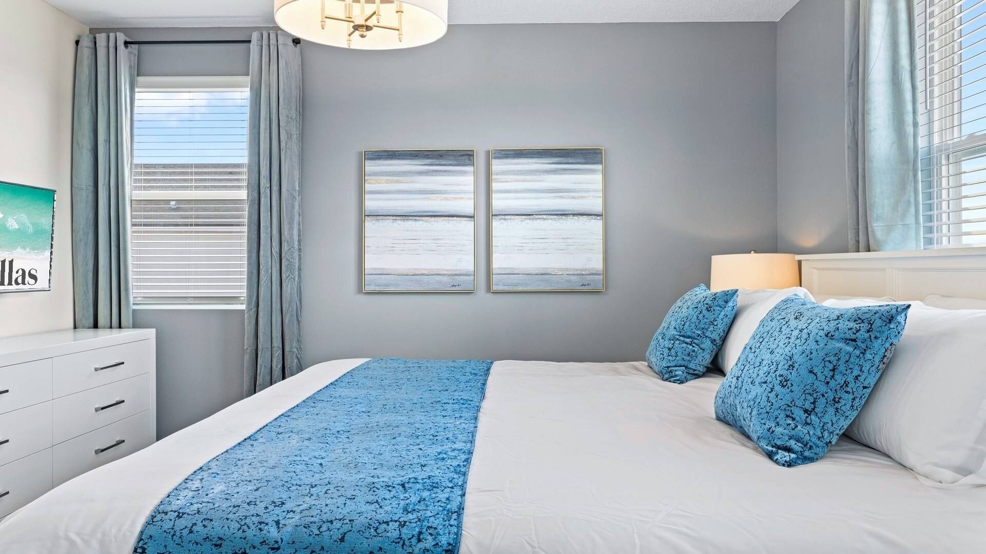 A bedroom with a bed, two wall-mounted paintings, a bedside lamp, and a window with curtains.