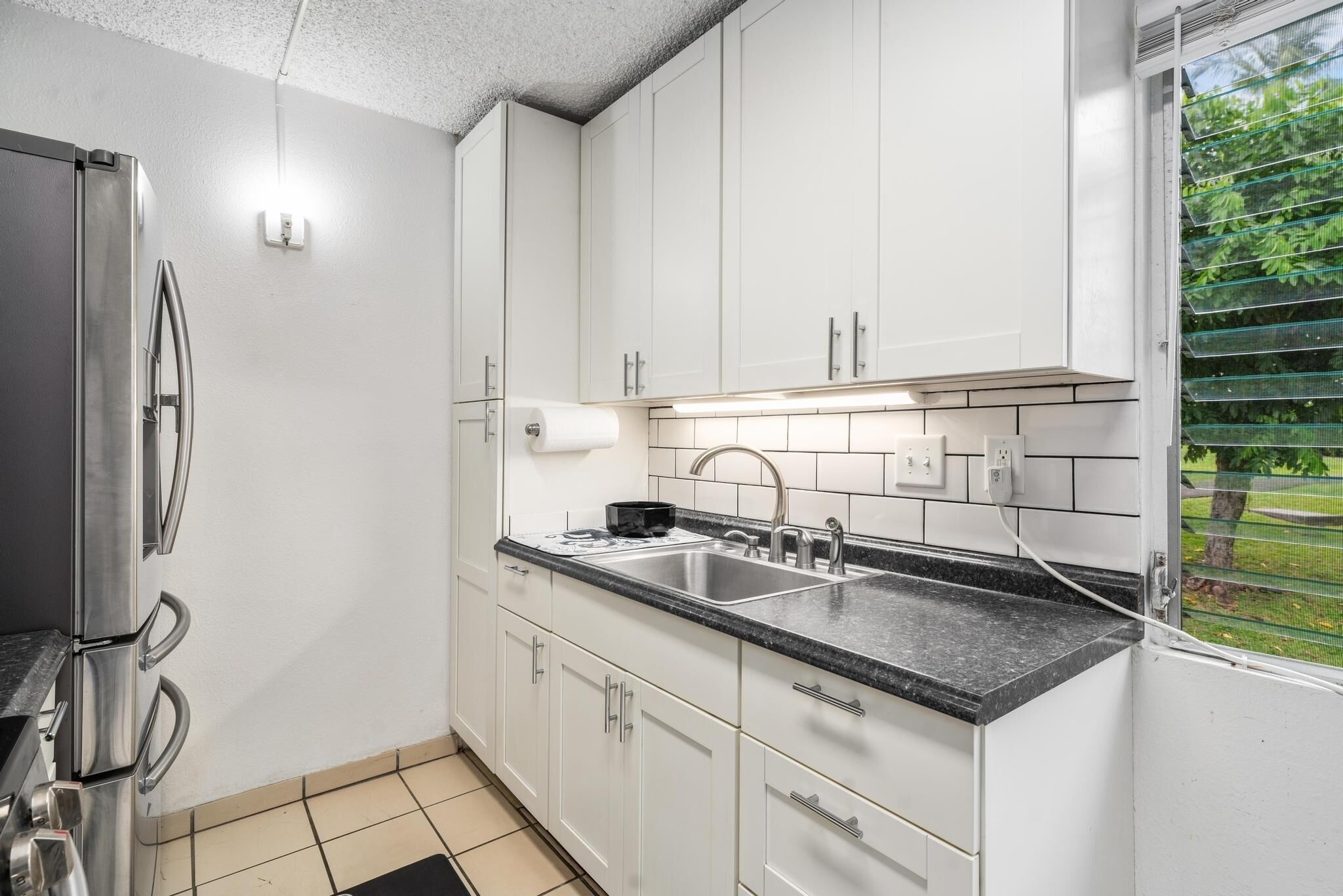 A modern kitchen with white cabinets, a stainless steel refrigerator, a black countertop, and a window with a view of greenery.