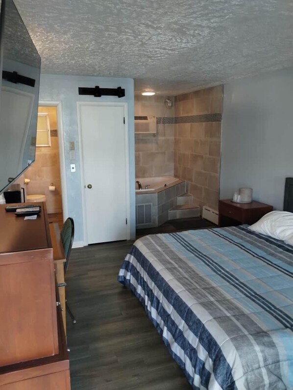 A bedroom with a bed, a desk, a chair, a bathroom with a shower, and a door. - Riviera Motel Erie (Erie)