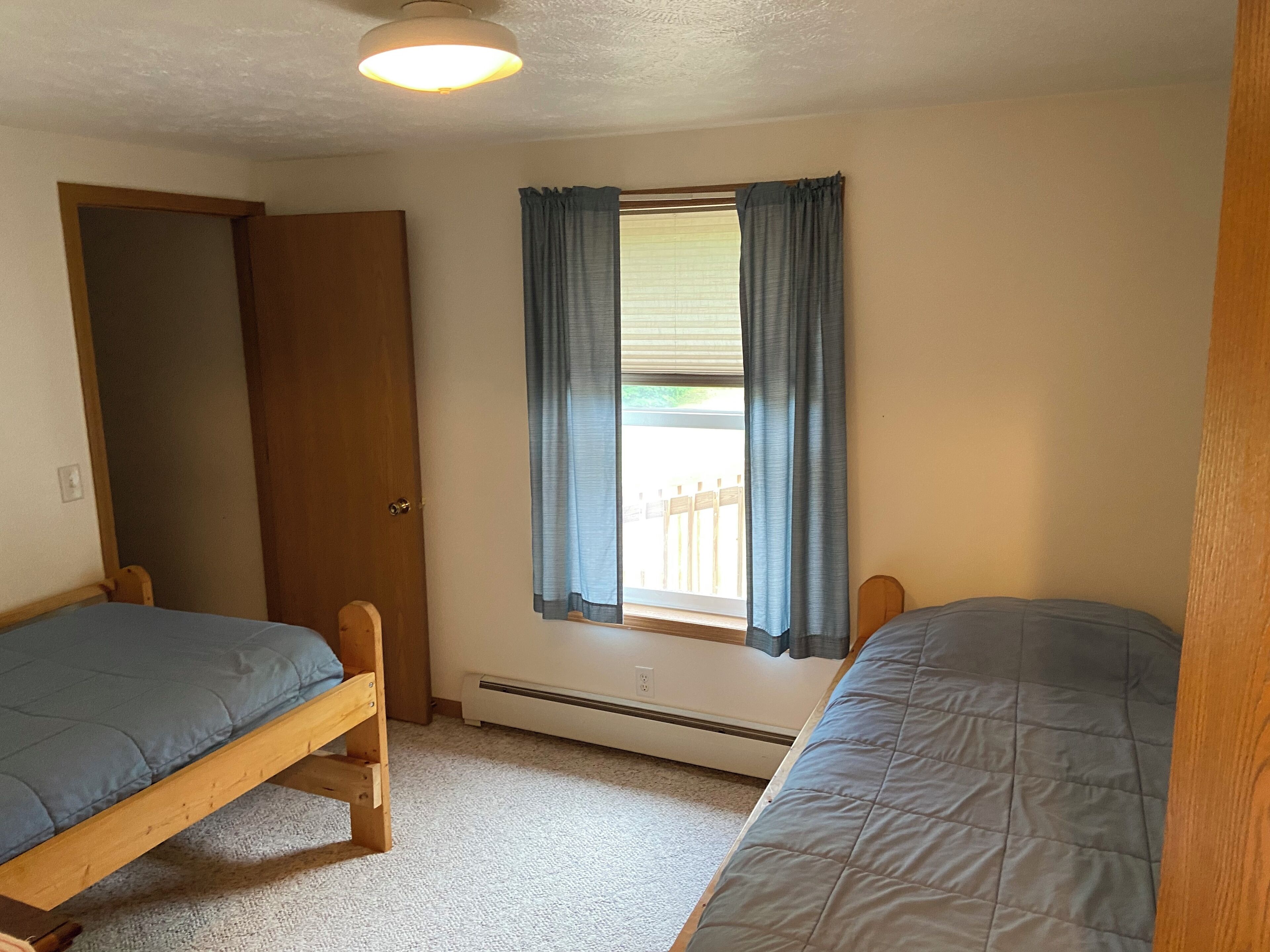 A room with two wooden beds, a window with blue curtains, a ceiling light, and a door.