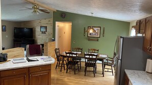 A kitchen with wooden cabinets, a marble countertop, a dining table with chairs, a refrigerator, and a television in the background. - Hickey Creek Retreat Secluded Log Home - 7 miles from Pictured Rocks Lake shore (Shingleton)