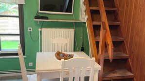 Une pièce avec un mur vert, une table blanche avec des chaises, un téléviseur fixé au-dessus de la table, un escalier en bois et une fenêtre avec des stores.