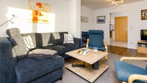 Un salon moderne avec un canapé gris, une table basse en bois et un fauteuil bleu. Un tableau est accroché au mur, et une porte donne accès à une autre pièce.
