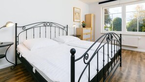 Une chambre à coucher avec un lit en métal noir, du linge de lit blanc, une table de chevet avec une lampe, une armoire en bois et une fenêtre donnant sur des arbres.