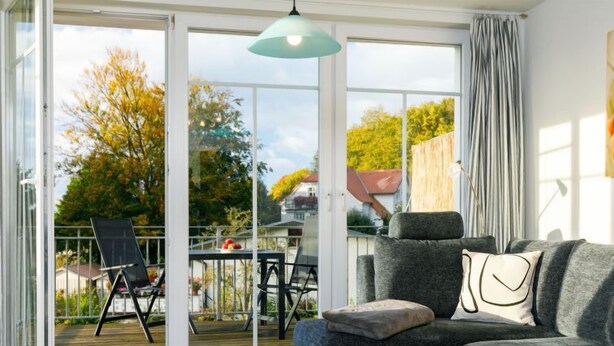 Un salon moderne avec un canapé gris, une table basse en bois et une porte vitrée donnant sur un balcon aménagé avec du mobilier d’extérieur.