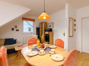 A dining area with a round table set for four, a wooden cabinet, and a window with a view of a blue building. - Apartment Martin by Interhome (Zinnowitz)