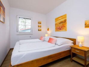 A double bed with white linens, a wooden nightstand with a lamp, and a window with a view of trees. - Apartment Jacobs by Interhome (Zinnowitz)