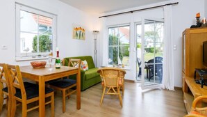 A spacious living area with a wooden dining table, green sofa, and wicker chairs. There is a sliding glass door leading to an outdoor patio. - Apartment Jacobs by Interhome (Zinnowitz)