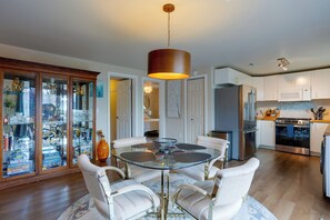 A modern kitchen with stainless steel appliances, a glass dining table with white upholstered chairs, and a wooden cabinet with glass doors.