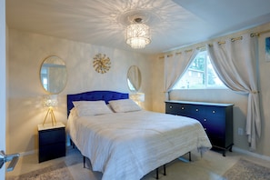 A bedroom with a bed, bedside tables, a dresser, a chandelier, a window with curtains, and wall decorations.