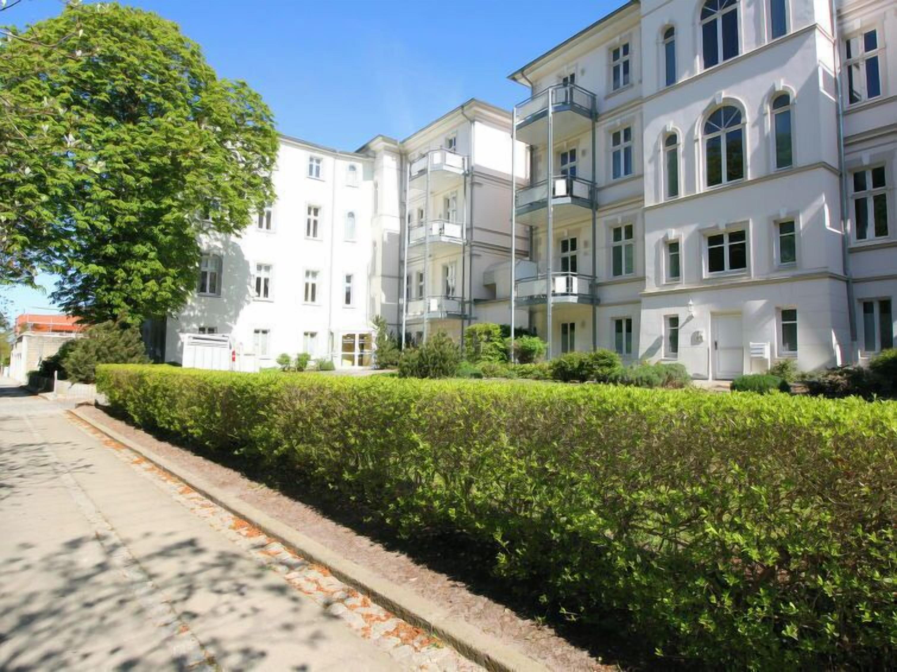 A row of white multi-story buildings with balconies, a clear sky, and a well-maintained hedge along the sidewalk.