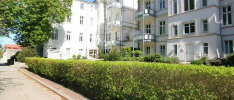 A row of white multi-story buildings with balconies, a clear sky, and a well-maintained hedge along the sidewalk.