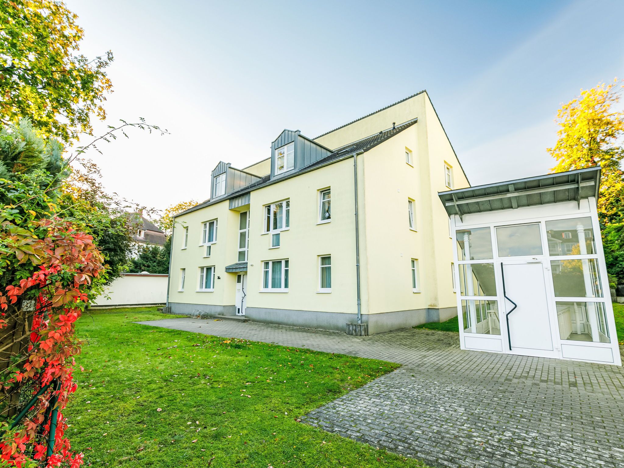 Un bâtiment à deux étages avec une façade jaune clair, une petite structure attenante dotée d’une porte en verre, et une cour pavée avec de l’herbe et un chemin en briques.