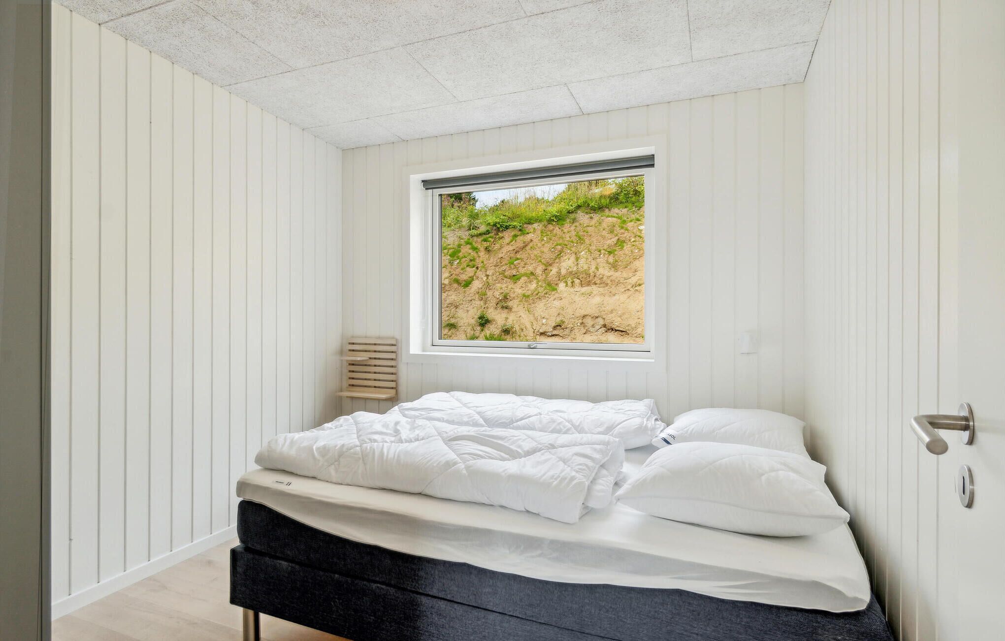 A bedroom with a bed, a window with a view of a beach, and white walls.