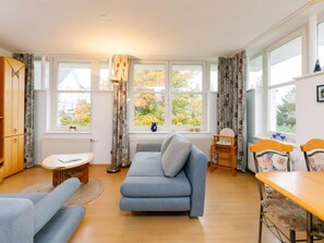 Una sala de estar acogedora con un sofá, una mesita y un comedor con sillas. Amplias ventanas ofrecen vistas al exterior.