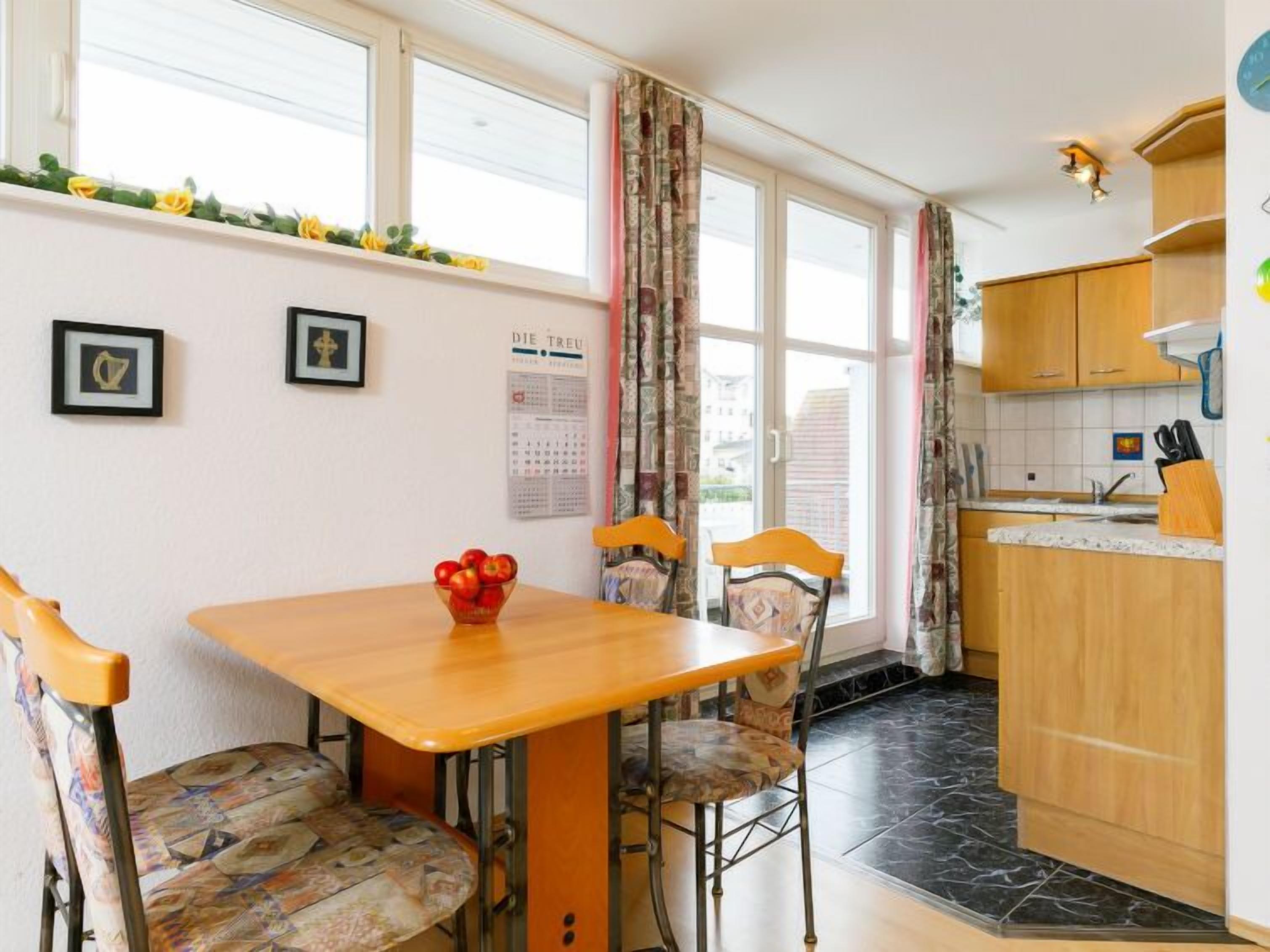 A small kitchen with a wooden table and chairs, a calendar on the wall, and a window with curtains.