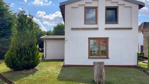 Ein zweistöckiges Haus mit weißer Fassade, einer Veranda und einem kleinen Vorgarten mit Baum und Holzmast.
