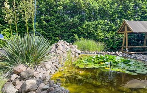 Um lago com pedras e nenúfares, rodeado por vegetação e uma estrutura de madeira.