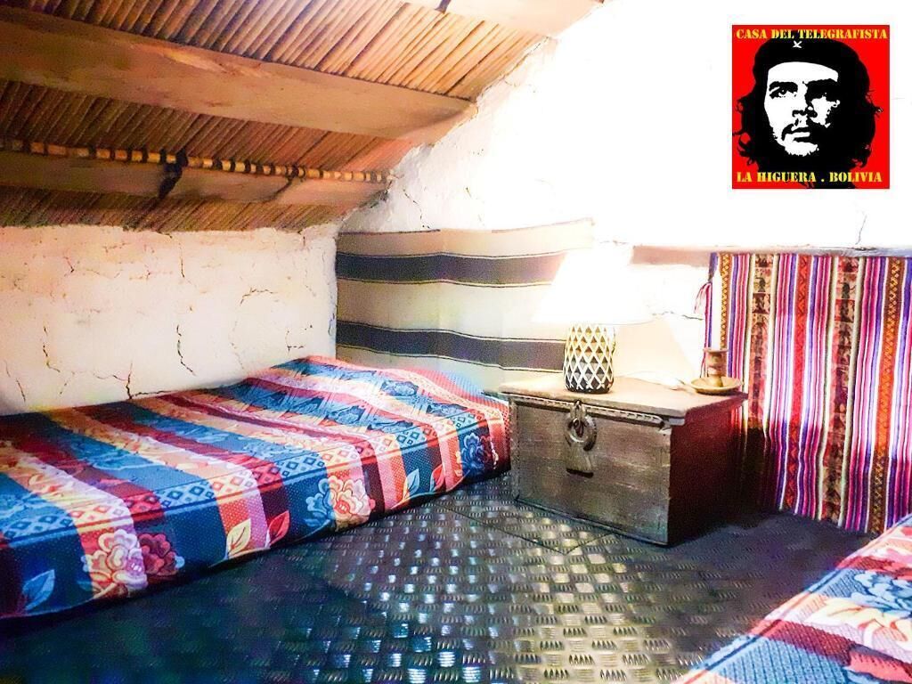 Um quarto pequeno e rústico, com teto inclinado, uma cama de madeira, um pequeno aparador e uma cortina colorida.