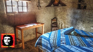 Ein rustikaler Raum mit einem Holztisch, einem Bett mit gemusterten Textilien, einem Fenster mit Ausblick und einem Regal mit einer Topfpflanze.