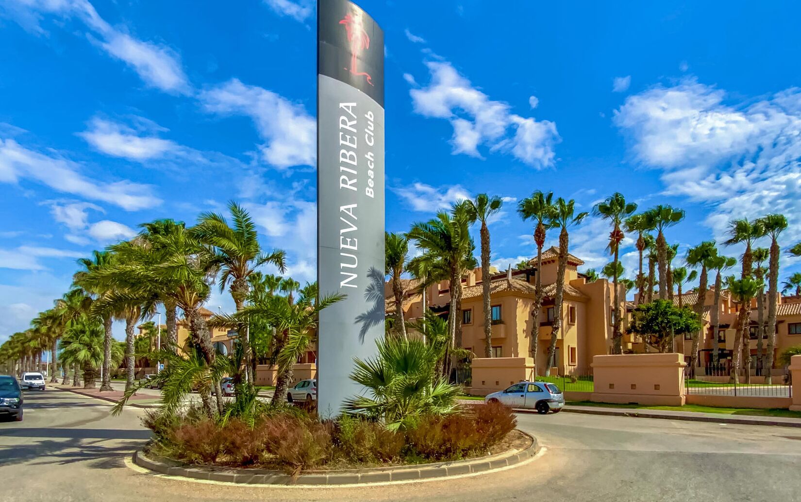 A tall sign with "NUEVA RIBERA Beach Club" written on it, surrounded by palm trees and a building with a tiled roof.