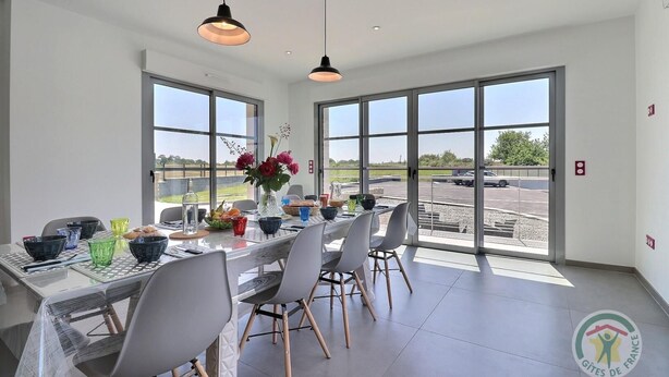 A dining area with a table set for six, large windows, and a view of a parking lot.