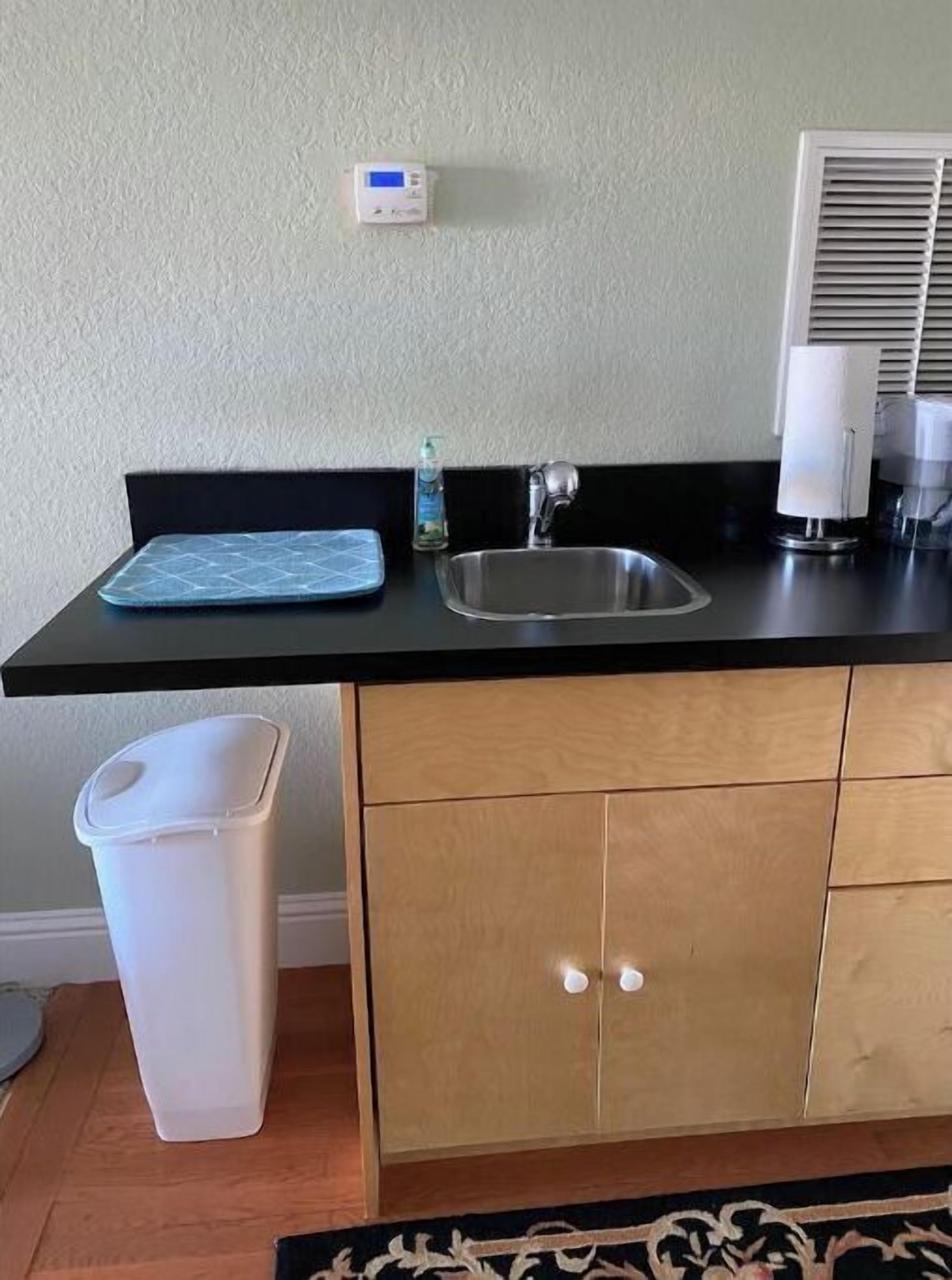 Una cocina con encimera negra, pileta de acero inoxidable y un cubo de basura blanco.