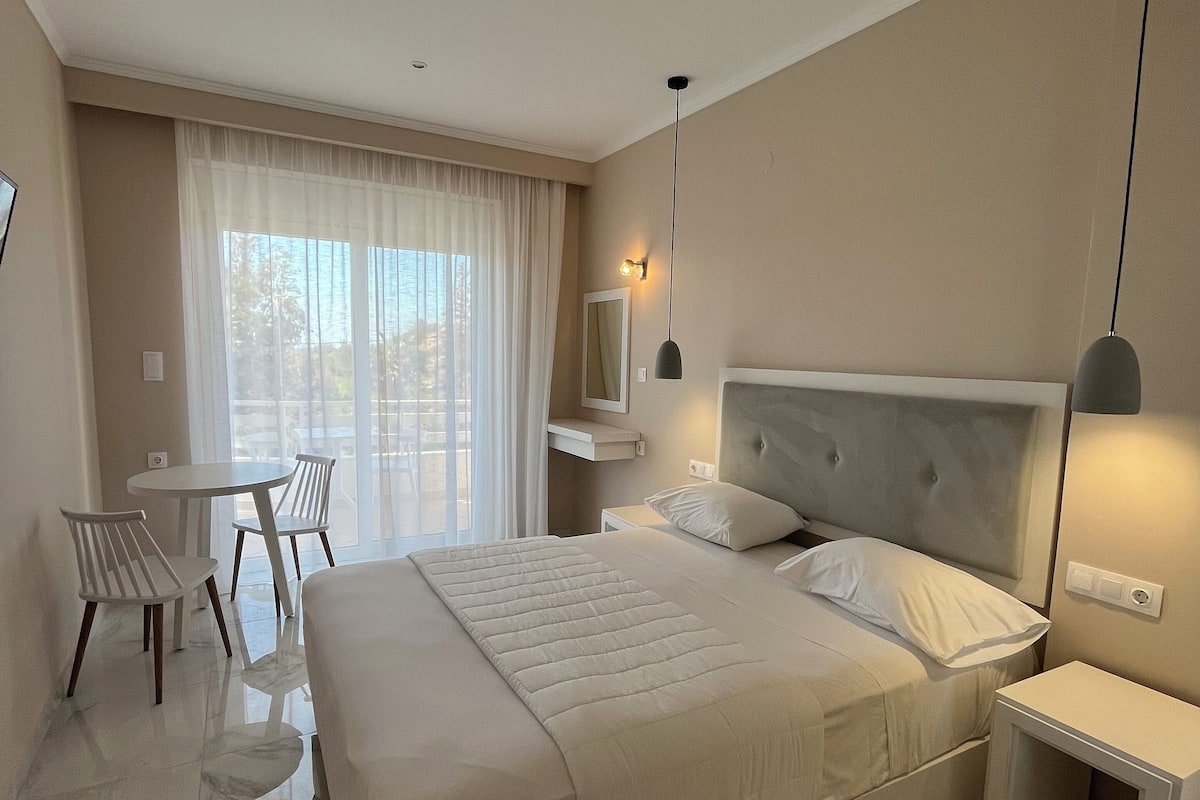 Ein modernes Hotelzimmer mit einem großen Bett, Nachttischen, einem Schreibtisch mit Stühlen und einem großen Fenster mit leichtem Vorhang.