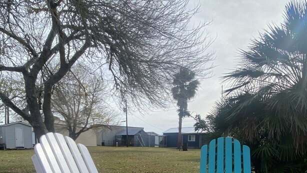 Two Adirondack chairs on a gravel path with a backdrop of trees and a building.
