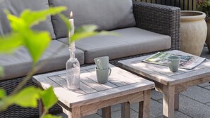 A cozy outdoor seating area with a wicker sofa, two wooden tables, a candle, and a vase.