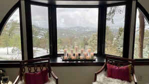 Ein Zimmer mit Erkerfenster mit Blick auf eine verschneite Landschaft, zwei Sessel mit flauschigen weißen und rosa Kissen sowie eine Ablage mit Kerzen am Fensterbrett.