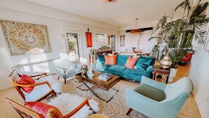 Ein großzügiger Wohnraum mit einem türkisfarbenen Sofa, Holzstühlen und einem Couchtisch. An den Wänden befinden sich Pflanzen und Kunstwerke, und ein großes Fenster lässt viel natürliches Licht herein.