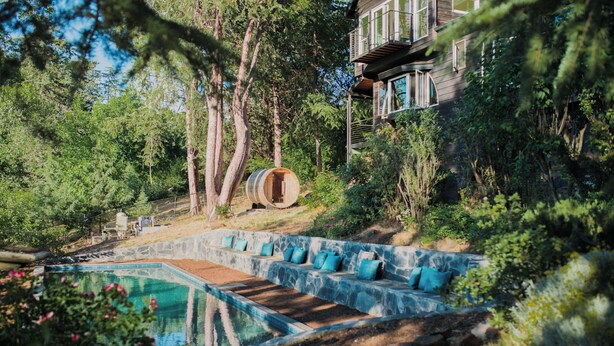 Ein Pool mit Steinkante und blauen Kissen, umgeben von Bäumen und einem Holzfass.