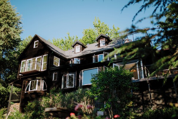 Une maison en bois à plusieurs étages, dotée de grandes fenêtres et d’un balcon, entourée d’une végétation luxuriante.
