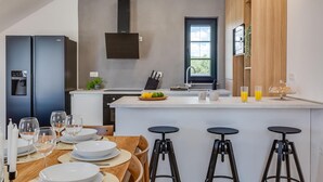 Een moderne keuken met een inbouwkoelkast, een waterdispenser en een raam met uitzicht op groen. Er is een eethoek met een tafel voor vier personen en drie barkrukken.