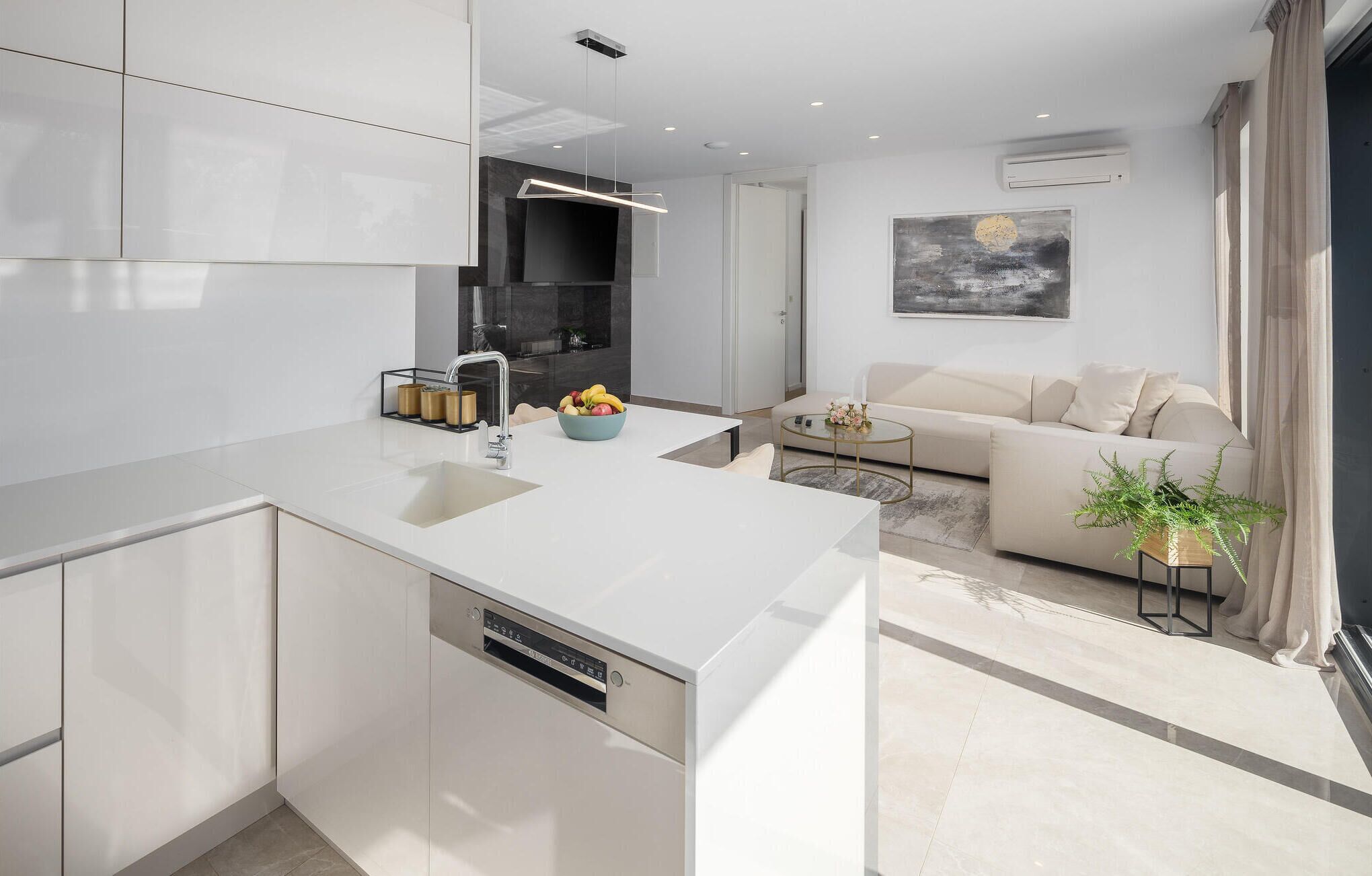 A modern kitchen with a white countertop, a built-in dishwasher, and a sink. There is a bowl of fruit on the counter and a small table with a vase of flowers.