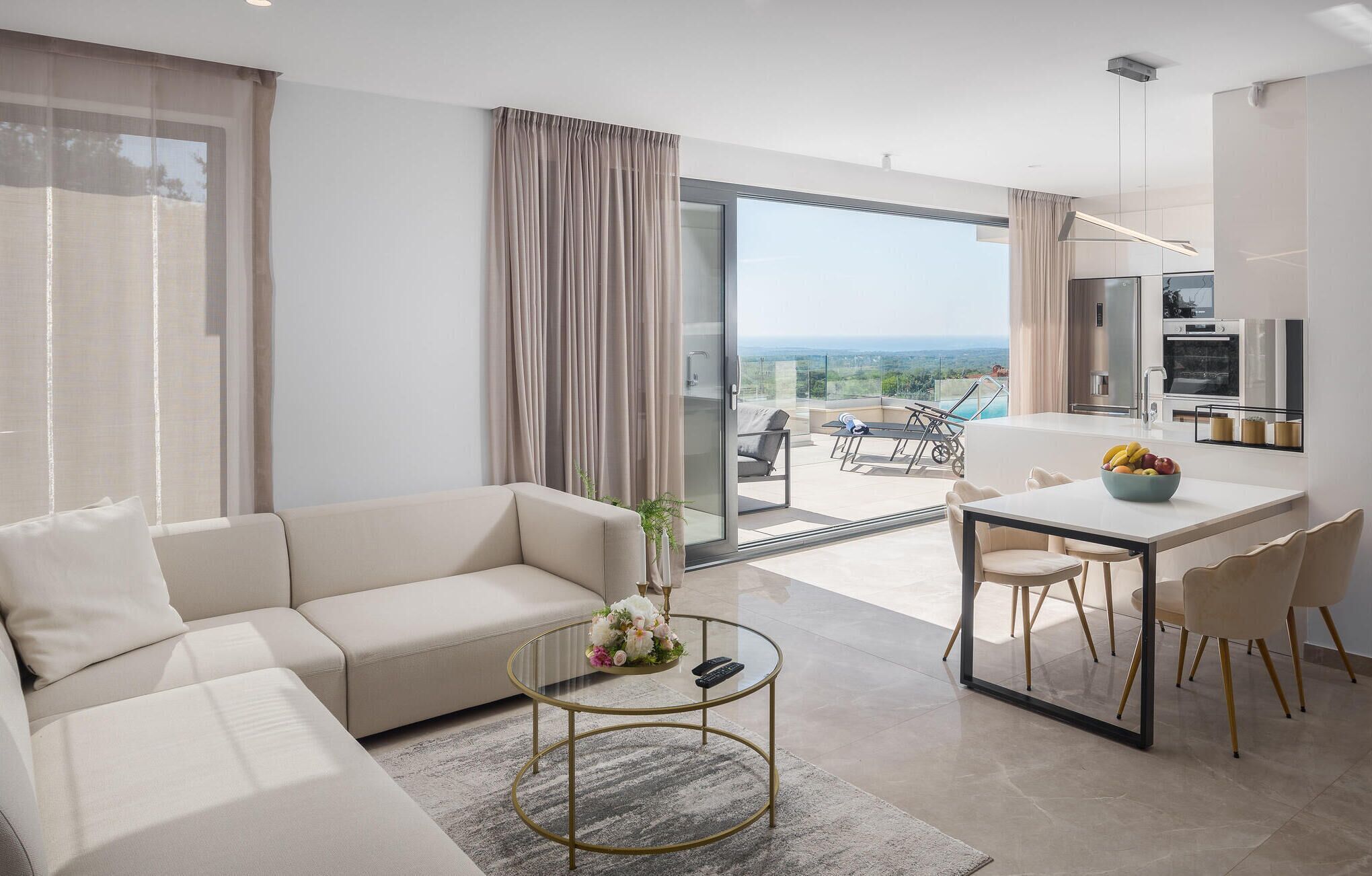 A modern living room with a sofa, coffee table, and dining area. A sliding glass door leads to a balcony with a view.