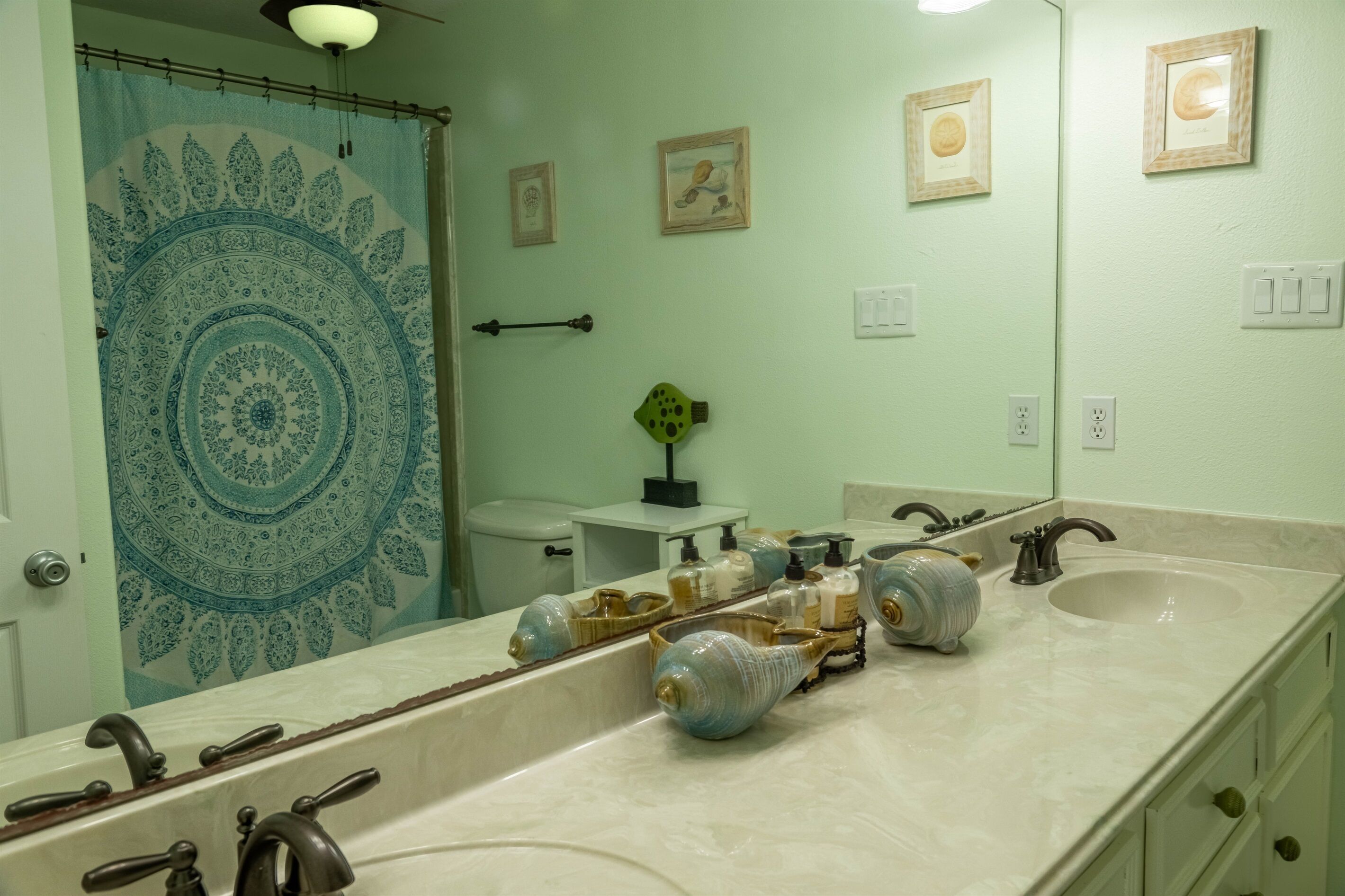 Un baño con un espejo grande, dos lavamanos y un inodoro. Hay elementos decorativos en el mostrador y un perchero para toallas.