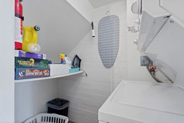 Ein kleines, kompaktes Badezimmer mit einem weißen Waschbecken, einem Spiegel und einem Duschbereich. Ein Regal beherbergt verschiedene Gegenstände, darunter einen Feuerlöscher, Reinigungsmittel und ein Brettspiel.