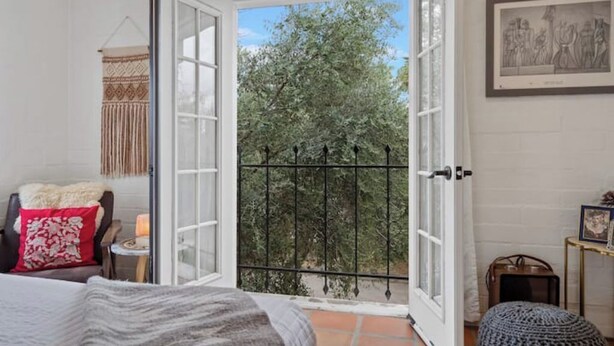 Une chambre avec une grande fenêtre, un lit, une chaise, une petite table et un tableau encadré au mur.