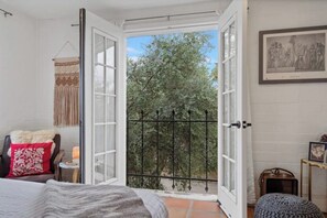 Een slaapkamer met een groot raam, een bed, een stoel, een klein tafeltje en een ingelijst schilderij aan de muur.
