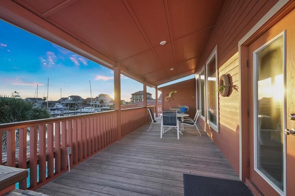 Une terrasse en bois avec une rambarde, une table entourée de chaises et, au loin, une vue sur des bateaux.