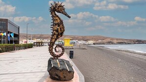 Une sculpture d’hippocampe sur un rocher, au bord de la plage, avec des bâtiments et un ciel dégagé en arrière-plan.