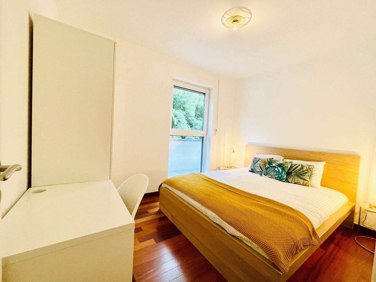 Un dormitorio con cama, escritorio y silla. Se ve follaje verde por la ventana.