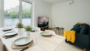 Ein modernes Wohnzimmer mit Esstisch für vier Personen, einer Couch, einem Fernseher und einem Balkon mit Blick auf Gebäude und Bäume.