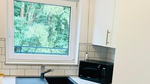 Eine kleine Küche mit weißen Schränken, einer schwarzen Mikrowelle, einem Fenster mit Blick auf Bäume und einem Spülbecken.