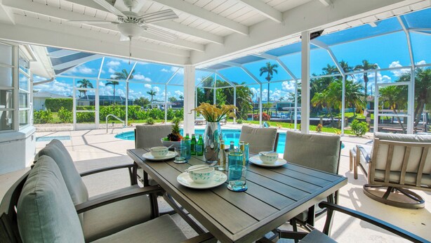 A covered outdoor dining area with a pool view, featuring a table set with plates, glasses, and a vase of flowers. - VRCC Villa Blue Pelican - Waterfront style & comfort (Cape Coral)