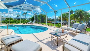 A pool area with a covered seating area, lounge chairs, and a table with a bottle and a mug. - VRCC Villa Blue Pelican - Waterfront style & comfort (Cape Coral)