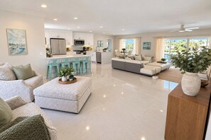 A modern kitchen with white cabinets, a central island with bar stools, a dining area with a sofa and chairs, and a living room with a large sectional sofa.